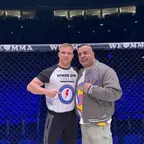 Zwei Männer im MMA-Käfig, einer zeigt auf sein Shirt vom Spikes Gym Berlin
